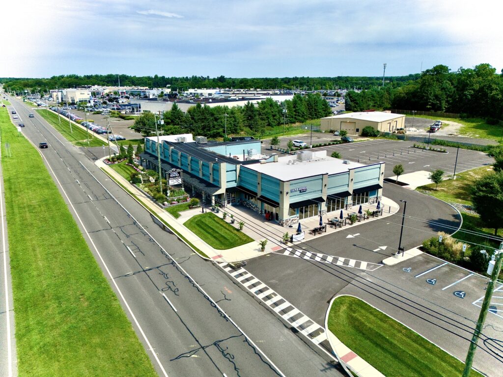 Aerial drone view of a modern commercial building with retail storefronts, outdoor seating, landscaped surroundings, and a large parking lot.
